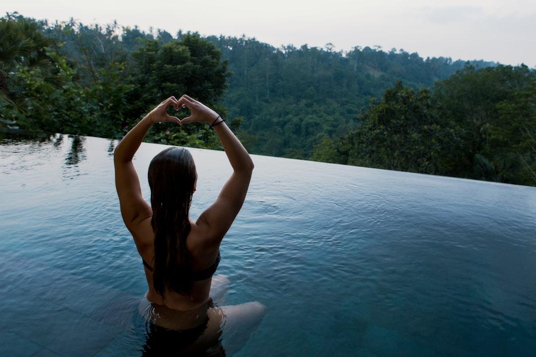 Woman in infinity pool making heart hand gesture facing green leafed trees from Jon Flobrant on Unsplash