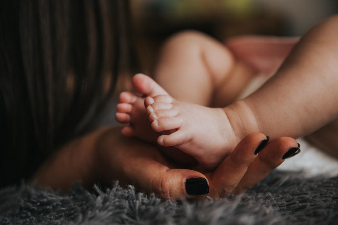 Infant's feet being held by a woman's hand with painted and manicured hands resting on a gray blanket from Alex Pasarelu on Unsplash